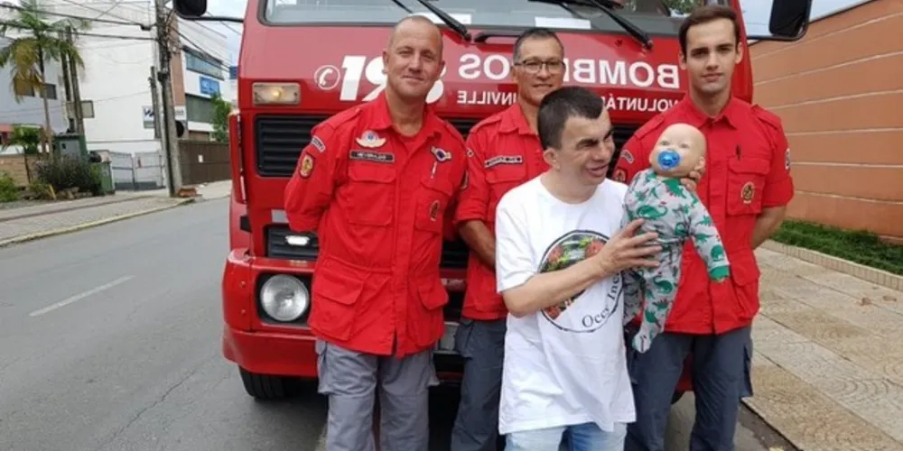 Sérgio da Silva segura o boneco Nico e posa para foto com bombeiros voluntários de Joinville
