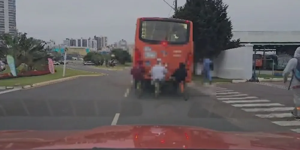 É possível ver três indivíduos montados em bicicletas enquanto seguram no veículo que transita pela via