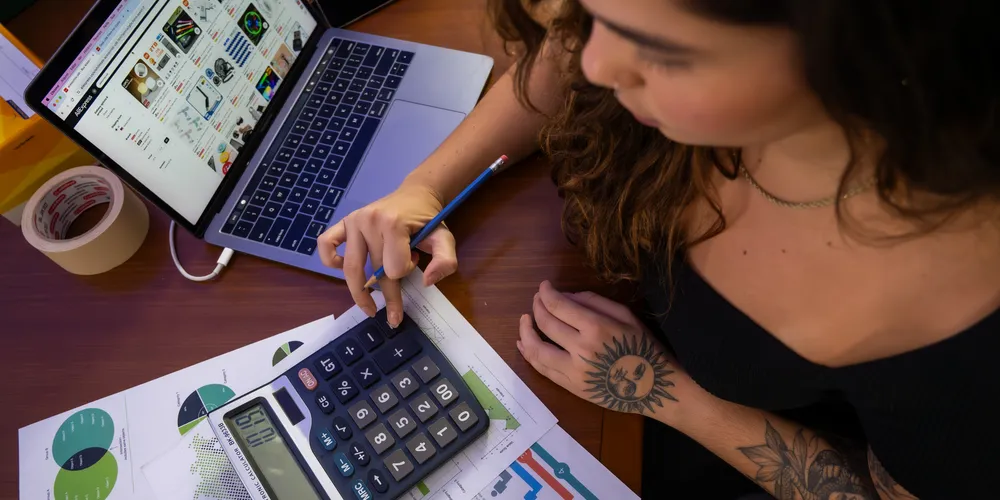 Mulher jovem trabalha em escritório com auxilio de calculadora e computador.