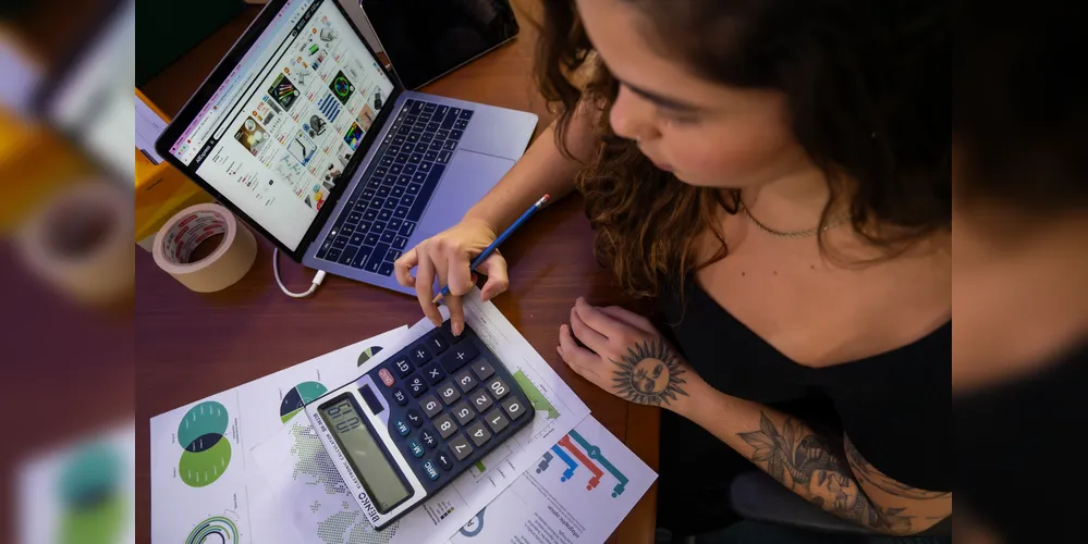 Mulher jovem trabalha em escritório com auxilio de calculadora e computador.