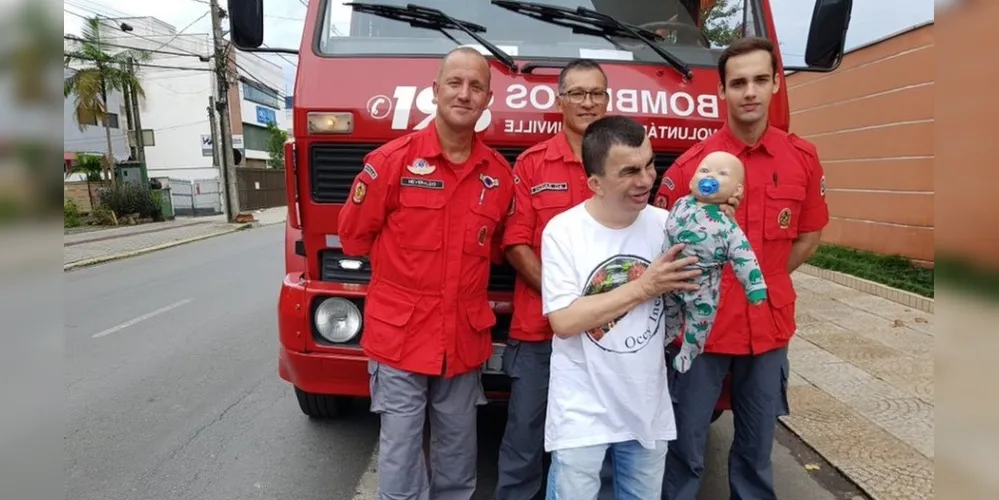 Sérgio da Silva segura o boneco Nico e posa para foto com bombeiros voluntários de Joinville