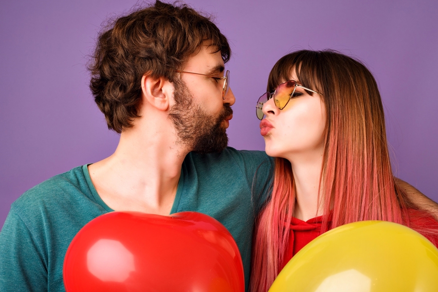 Close up studio portrait of happy hipster couple looking to each other and trying to kiss, holding balloons, bright trendy casual clothes and glasses, romantic mood.